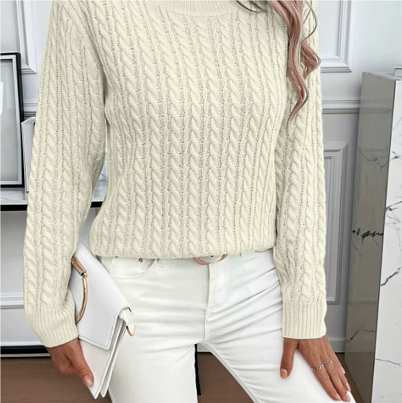 A woman wearing a cream-colored cable-knit sweater and white pants, holding a white handbag in an indoor setting.