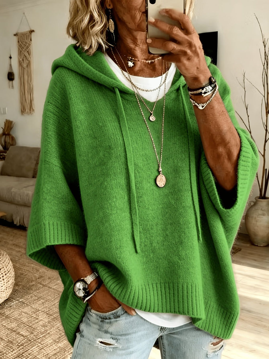 A woman takes a mirror selfie wearing a bright green knit poncho hoodie with drawstrings, layered over a white t-shirt and light-wash distressed jeans. She is accessorized with layered necklaces, bracelets, and a watch.