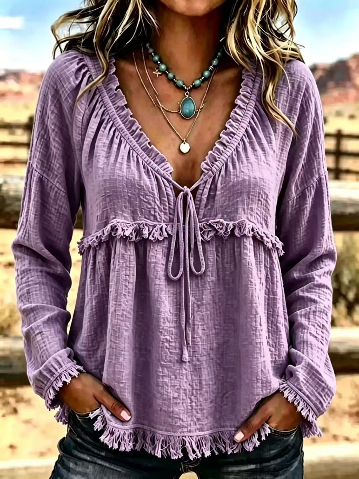 A woman wearing a long-sleeve lavender-purple crinkle gauze blouse with a ruffled V-neckline, tie-front detail, and frayed cuffs and hem, photographed outdoors against a blurred rustic landscape.