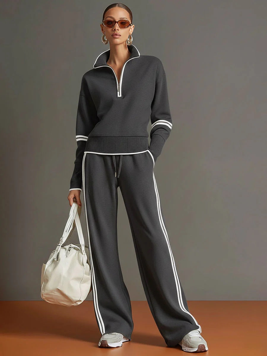 Woman wearing a dark gray tracksuit with white stripes, holding a white bag against a gray background.