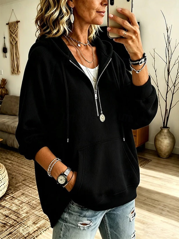 A woman takes a mirror selfie wearing a black zip-up hoodie with a front kangaroo pocket, layered over a white top and paired with distressed light-wash jeans. She is accessorized with layered necklaces, bracelets, and a watch.