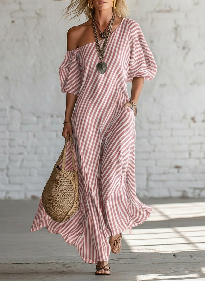 Woman wearing a pink and white striped dress with a woven bag against a white brick wall.