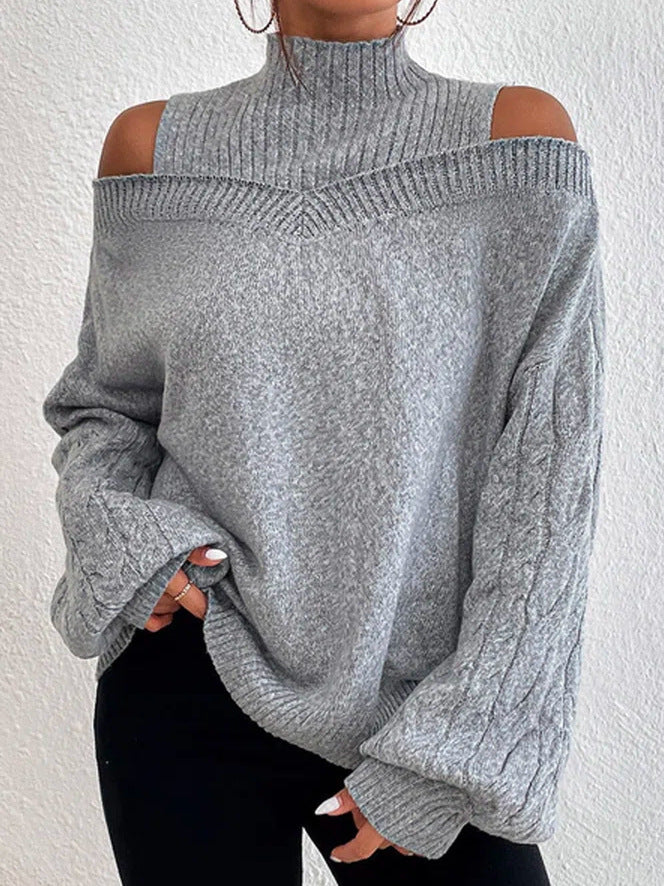 A person wearing a light gray ribbed knit sweater with cold-shoulder cutouts and a high mock neck, paired with black pants, standing against a white textured wall.
