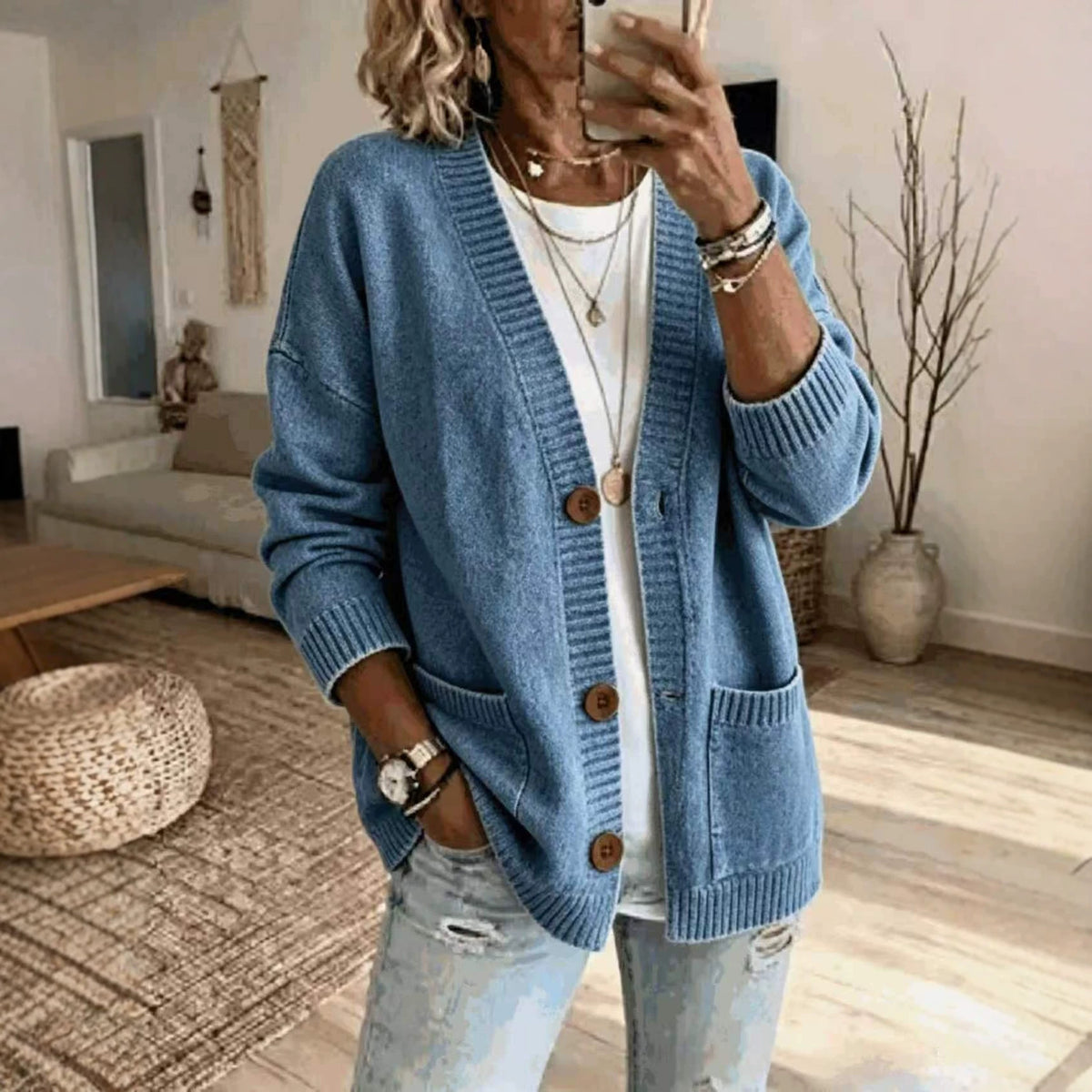A woman takes a mirror selfie wearing a light blue knit cardigan with brown wooden buttons over a white t-shirt and light-wash distressed jeans, in a cozy living room setting.