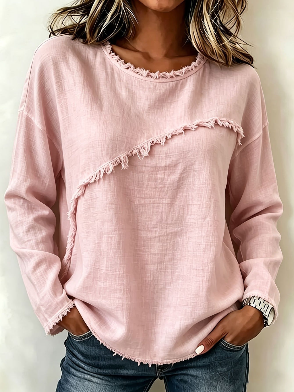 A woman wearing a light pink long-sleeve top with raw, frayed trim along the neckline and a diagonal frayed detail across the front, paired with blue jeans.