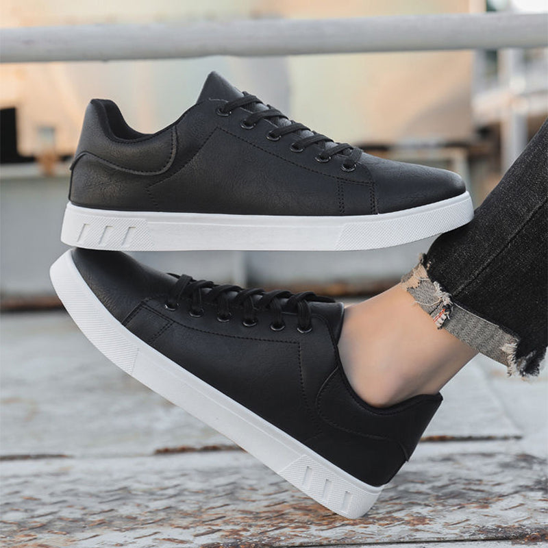 Black sneakers with white soles worn by a person on a wooden floor.