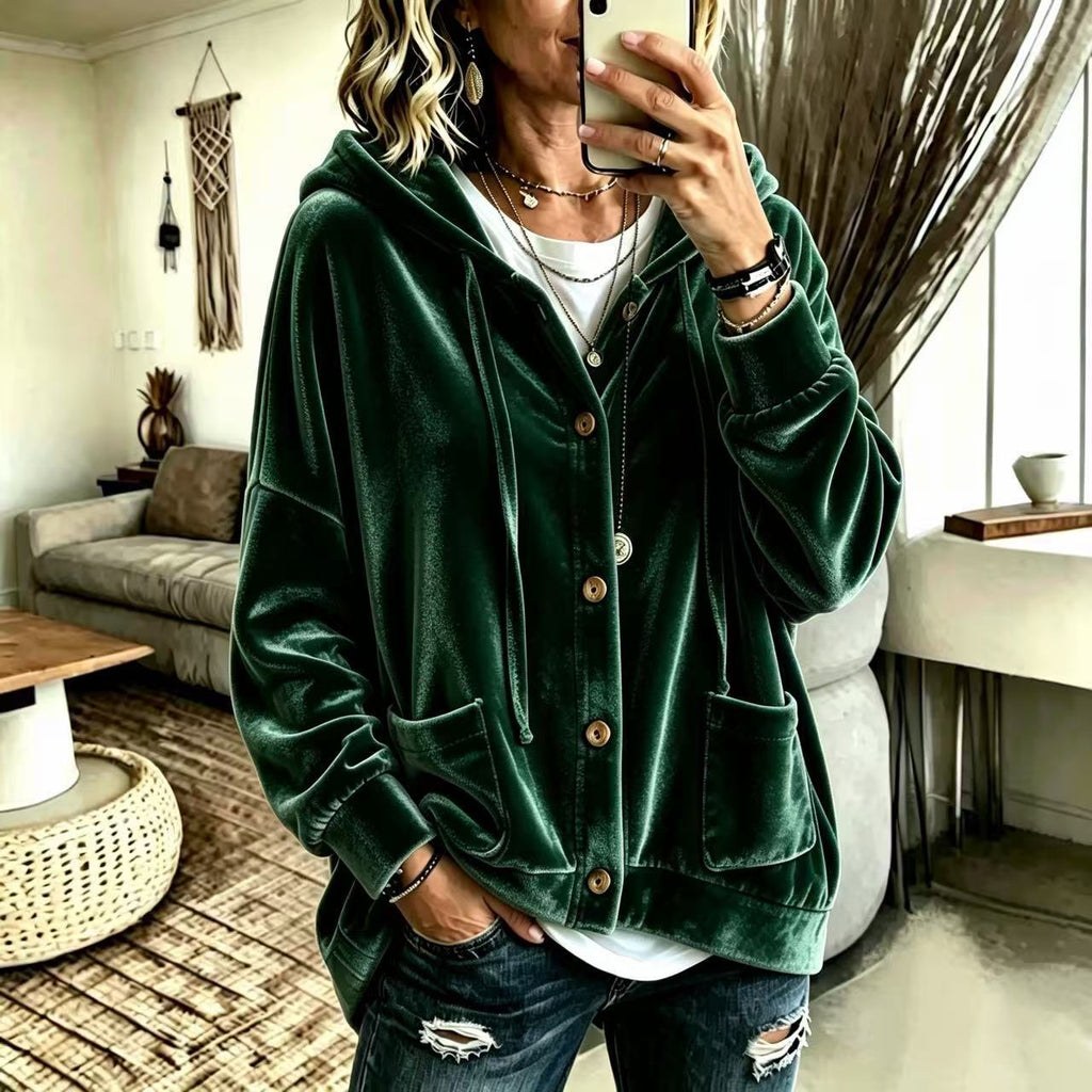 A woman takes a mirror selfie wearing a dark green velvet button-up hoodie jacket with a white t-shirt and distressed dark blue jeans in a modern living room setting.
