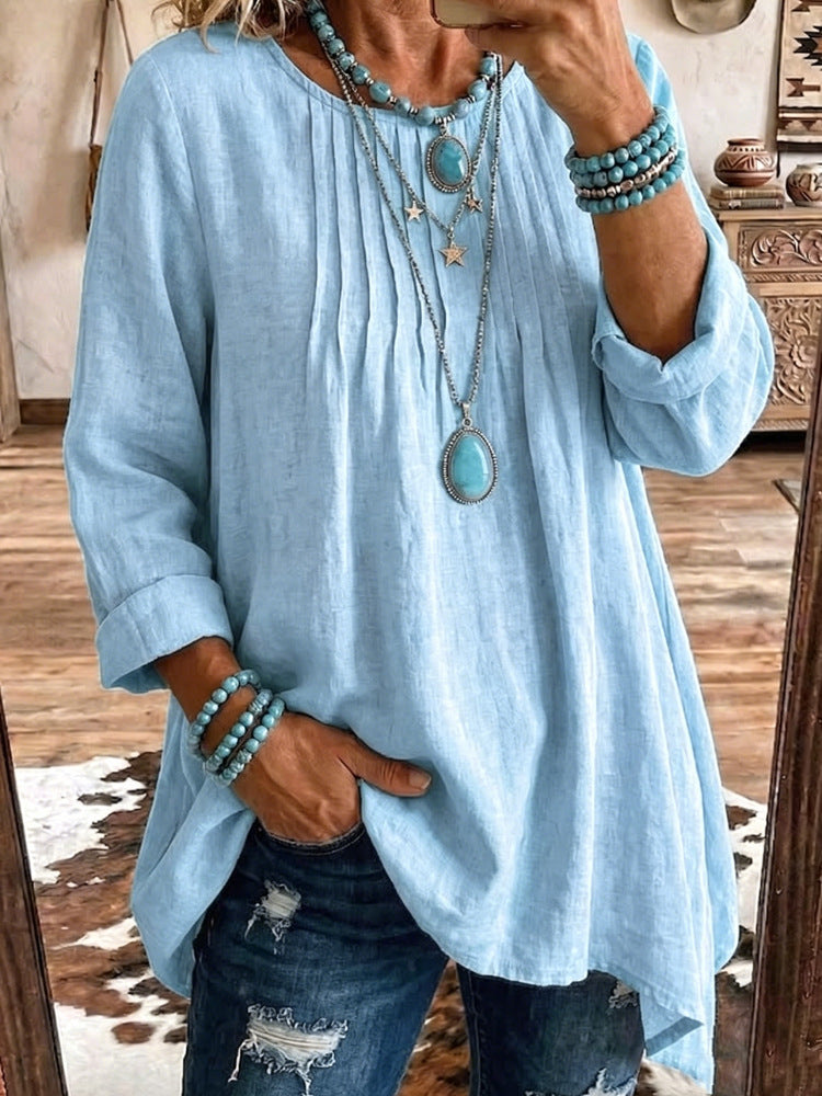 A woman takes a mirror selfie wearing a light blue linen-blend tunic top with rolled sleeves, layered silver necklaces, and turquoise bead bracelets, paired with distressed blue jeans.