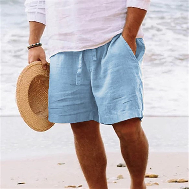 Person wearing light blue shorts and a white shirt on a beach, holding a straw hat.