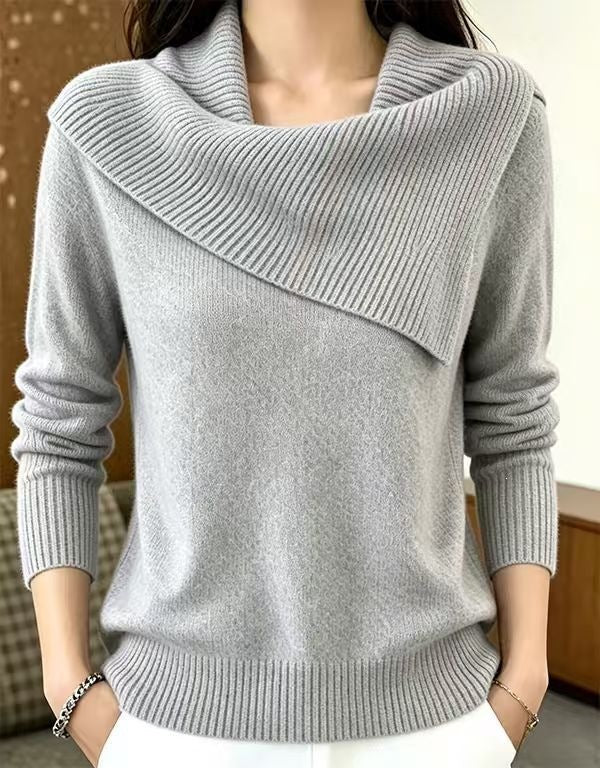 A woman wearing a light gray knit sweater with an asymmetrical draped cowl neck, ribbed cuffs, and a ribbed hem, paired with white pants.