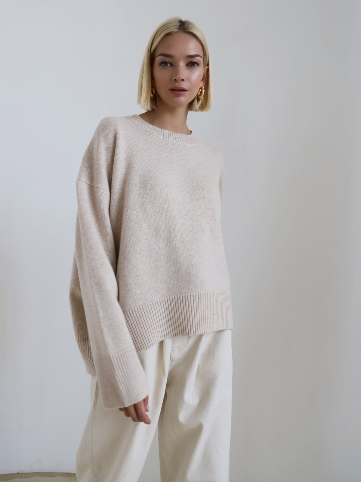 Woman wearing a beige sweater and white pants against a plain background