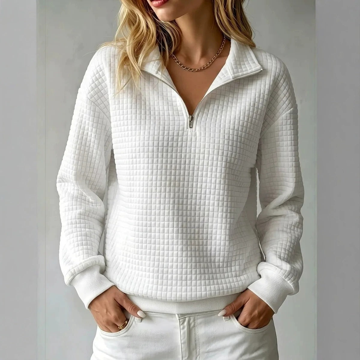A woman wearing a white textured quarter-zip pullover sweater with a stand collar, paired with white pants, shown from the lips down against a light gray background.