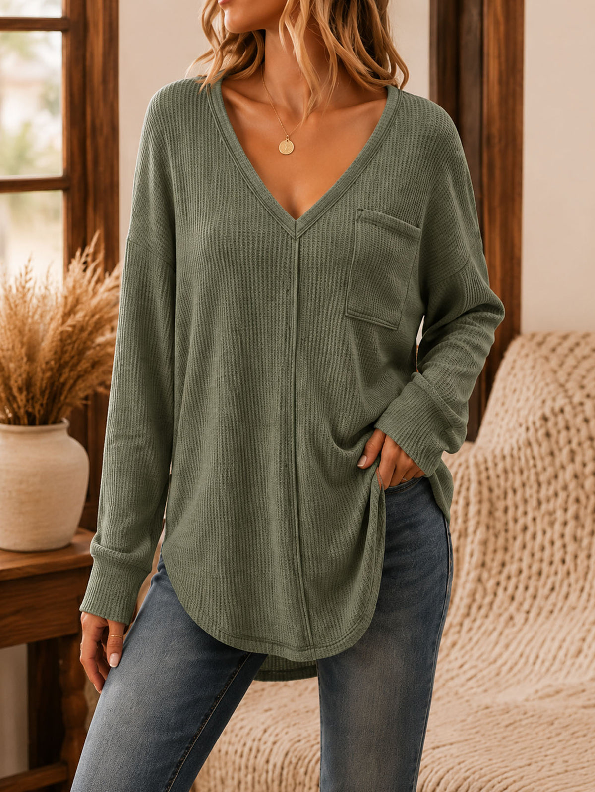A woman wearing a ribbed long-sleeve V-neck top in sage green with a chest pocket, paired with blue jeans, standing in a warm-toned indoor setting.
