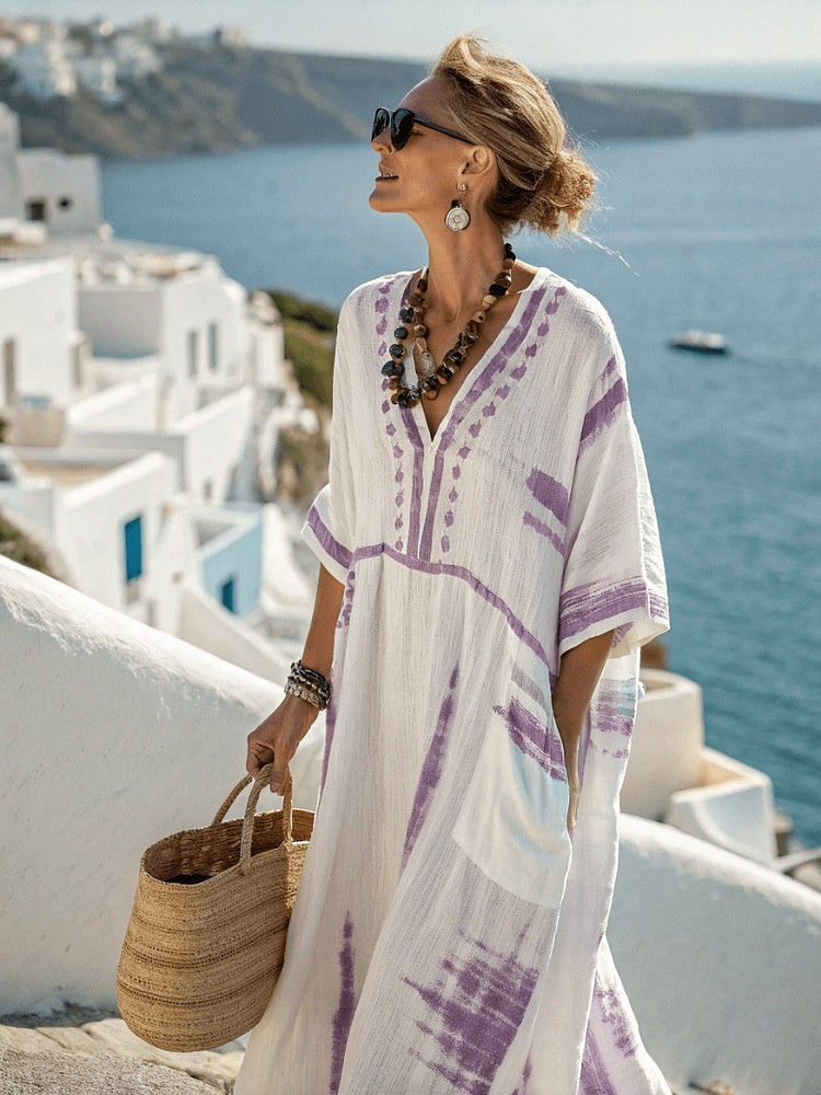 Woman in a white and purple dress with sunglasses and a necklace, standing on a scenic overlook with ocean and buildings in the background.