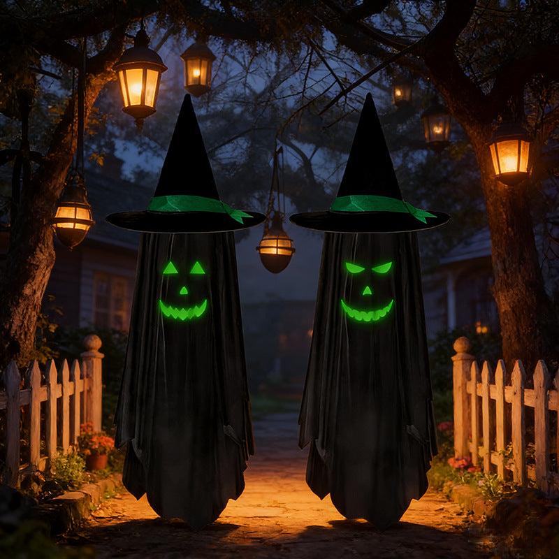 Two witch-like figures in black hooded cloaks with glowing green faces stand on a Halloween-lit path between trees and lanterns.