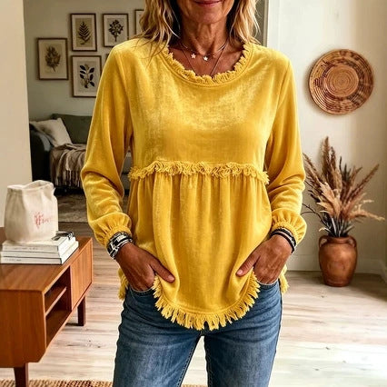 A woman wearing a yellow long-sleeve top with a ruffled neckline, a fringed waist seam, and a fringed hem, paired with blue jeans in a cozy indoor setting.