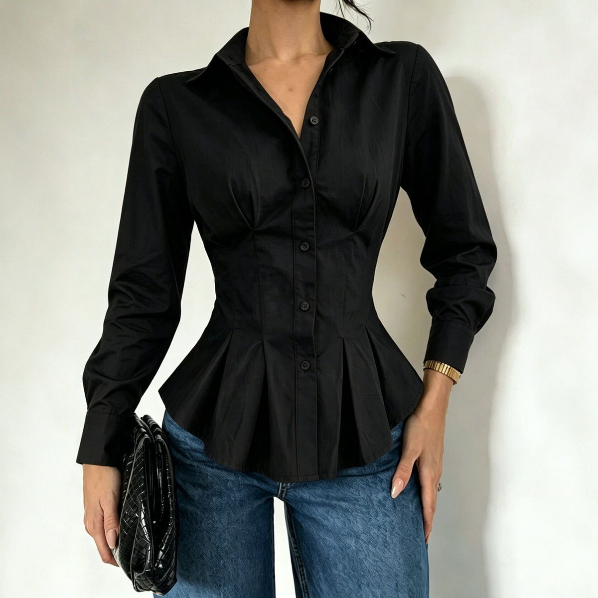 A woman wearing a fitted black long-sleeve button-front blouse with a peplum waist, paired with blue jeans, holding a black clutch against a plain light background.