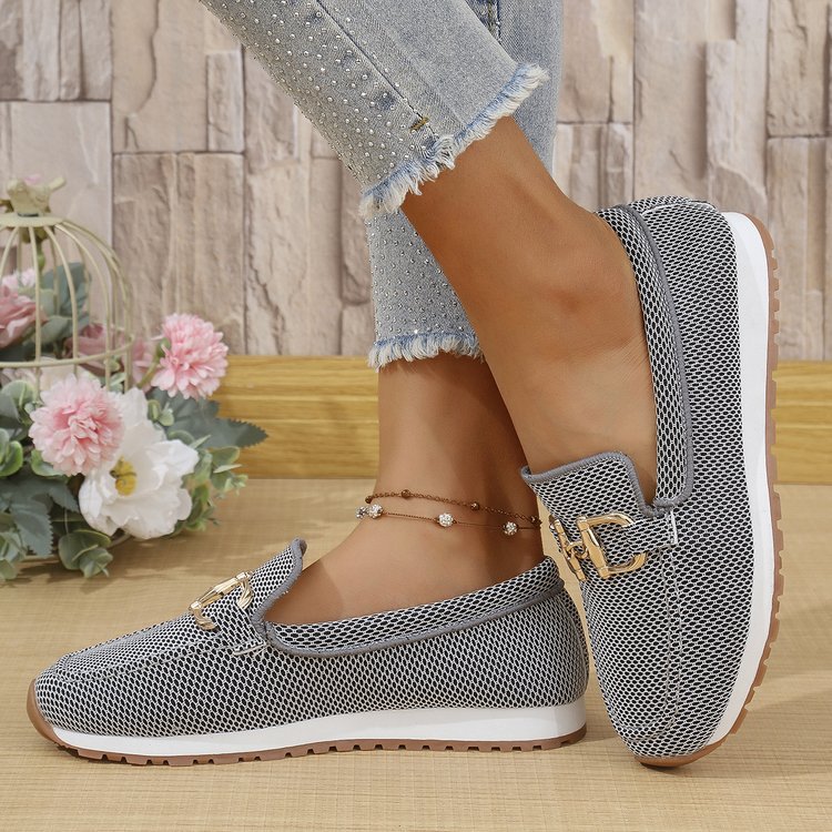 Gray patterned loafers worn with light blue jeans on a wooden floor.