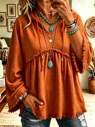 A woman wearing a rust-orange hooded tunic top with drawstrings, fringe trim, and wide three-quarter sleeves, paired with distressed blue jeans and layered turquoise and silver necklaces in a home setting.