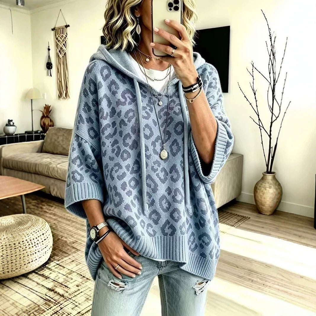 A woman takes a mirror selfie in a cozy living room, wearing an oversized light blue leopard-print hooded sweater with wide three-quarter sleeves and ribbed cuffs and hem.
