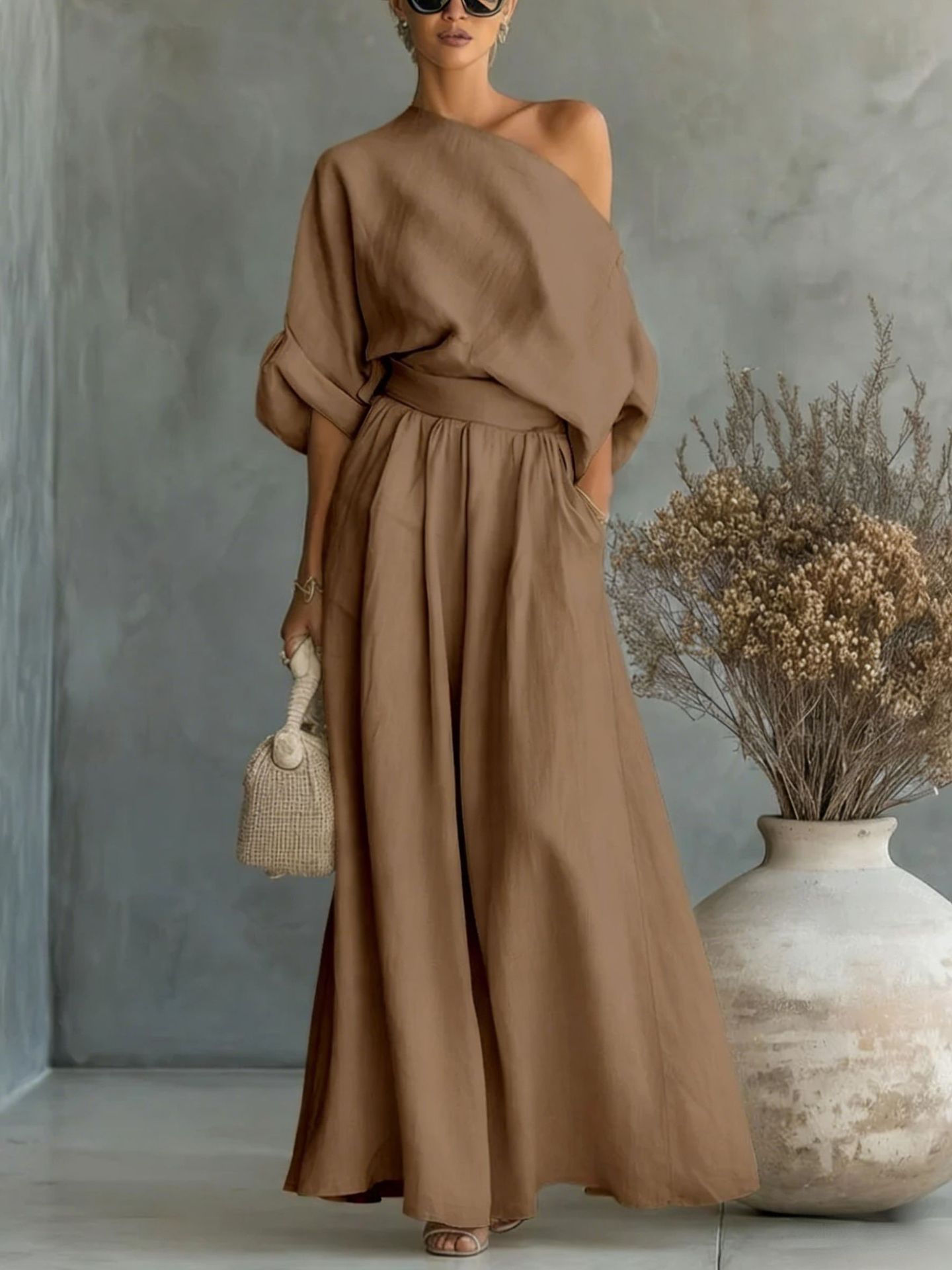 Woman wearing a brown off-shoulder dress holding a woven bag against a gray wall with decorative elements.