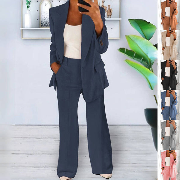 Person wearing a navy blue suit with a white top, standing in a room with a plant and shelves.