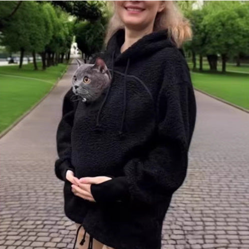 A woman wearing a black fuzzy hoodie with a gray cat inside the pocket, standing on a paved path in a park.