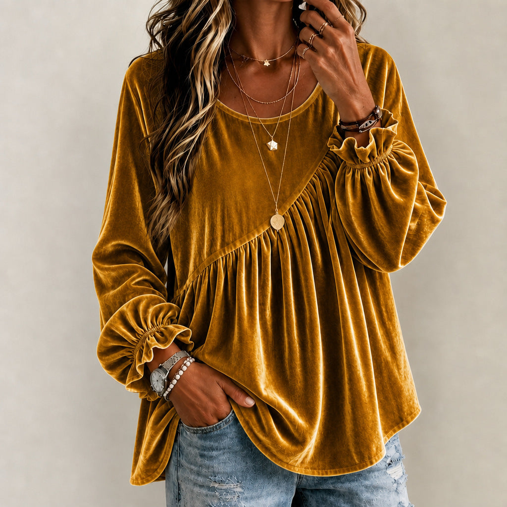A woman wearing a mustard yellow velvet long-sleeve blouse with a gathered peplum-style waist and ruffled cuffs, paired with light blue jeans and layered gold necklaces, against a neutral gray background.