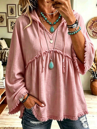A woman wearing a dusty pink hooded tunic top with drawstrings, a gathered waist, and frayed hems, paired with distressed blue jeans and turquoise statement jewelry.