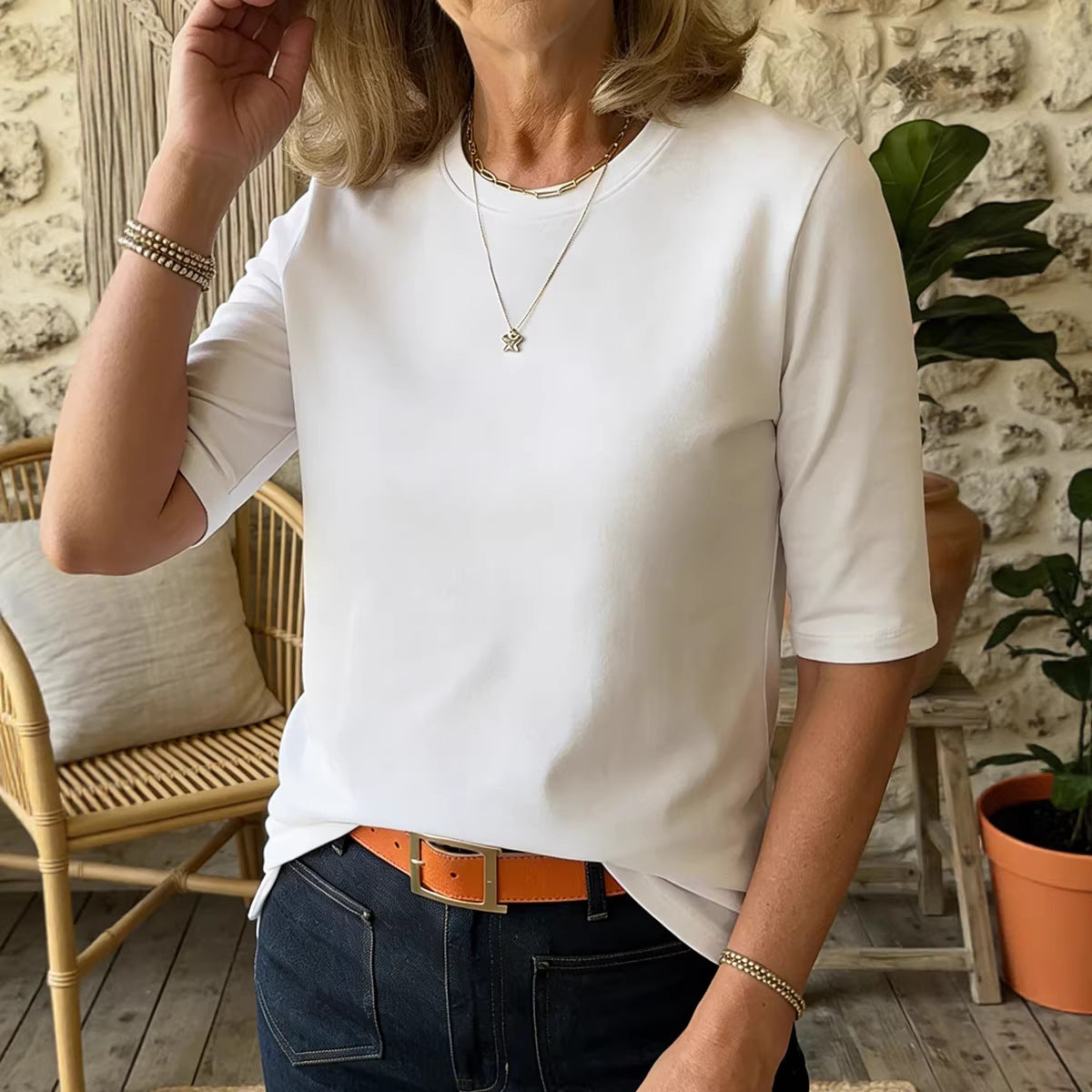 A woman wearing a white short-sleeve crewneck t-shirt, styled with dark blue jeans, an orange belt, and layered gold necklaces, standing in a rustic indoor setting with a stone wall and plants.