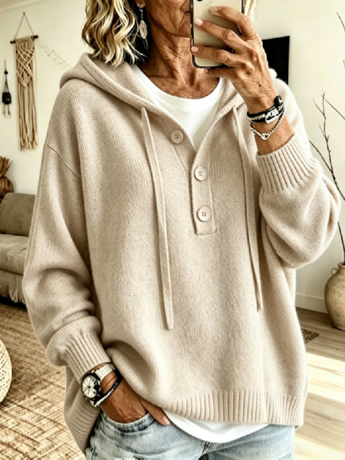 A woman takes a mirror selfie wearing a beige knit hooded sweater with a three-button placket over a white t-shirt and light-wash jeans, in a cozy living room setting.