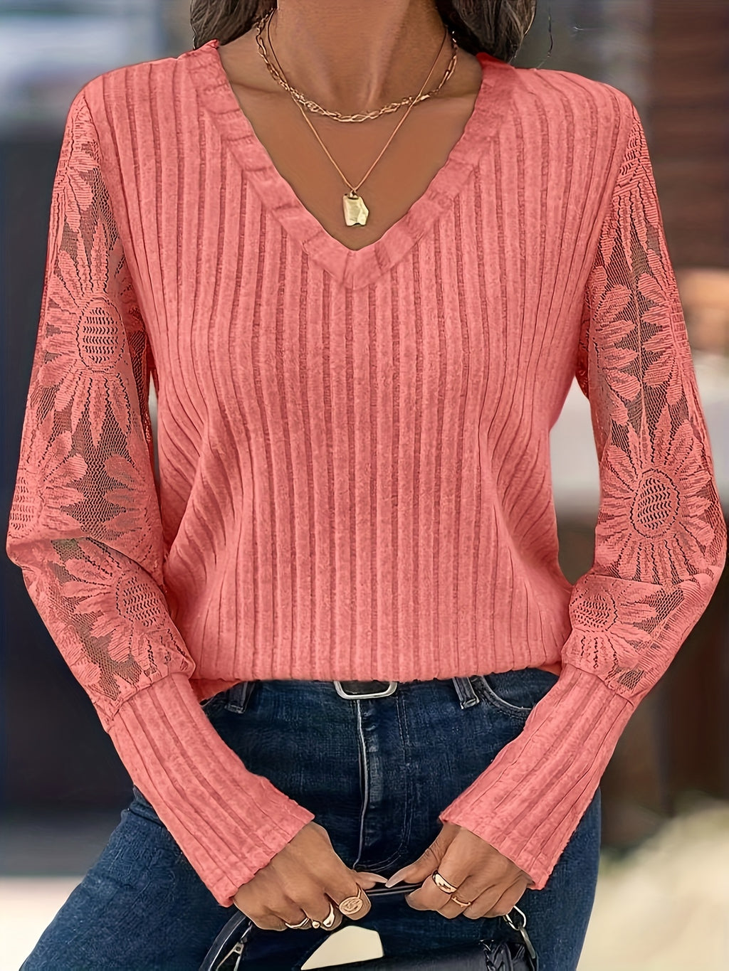 A woman wearing a coral pink ribbed knit sweater with sheer sunflower-patterned long sleeves, paired with blue jeans and layered gold necklaces.