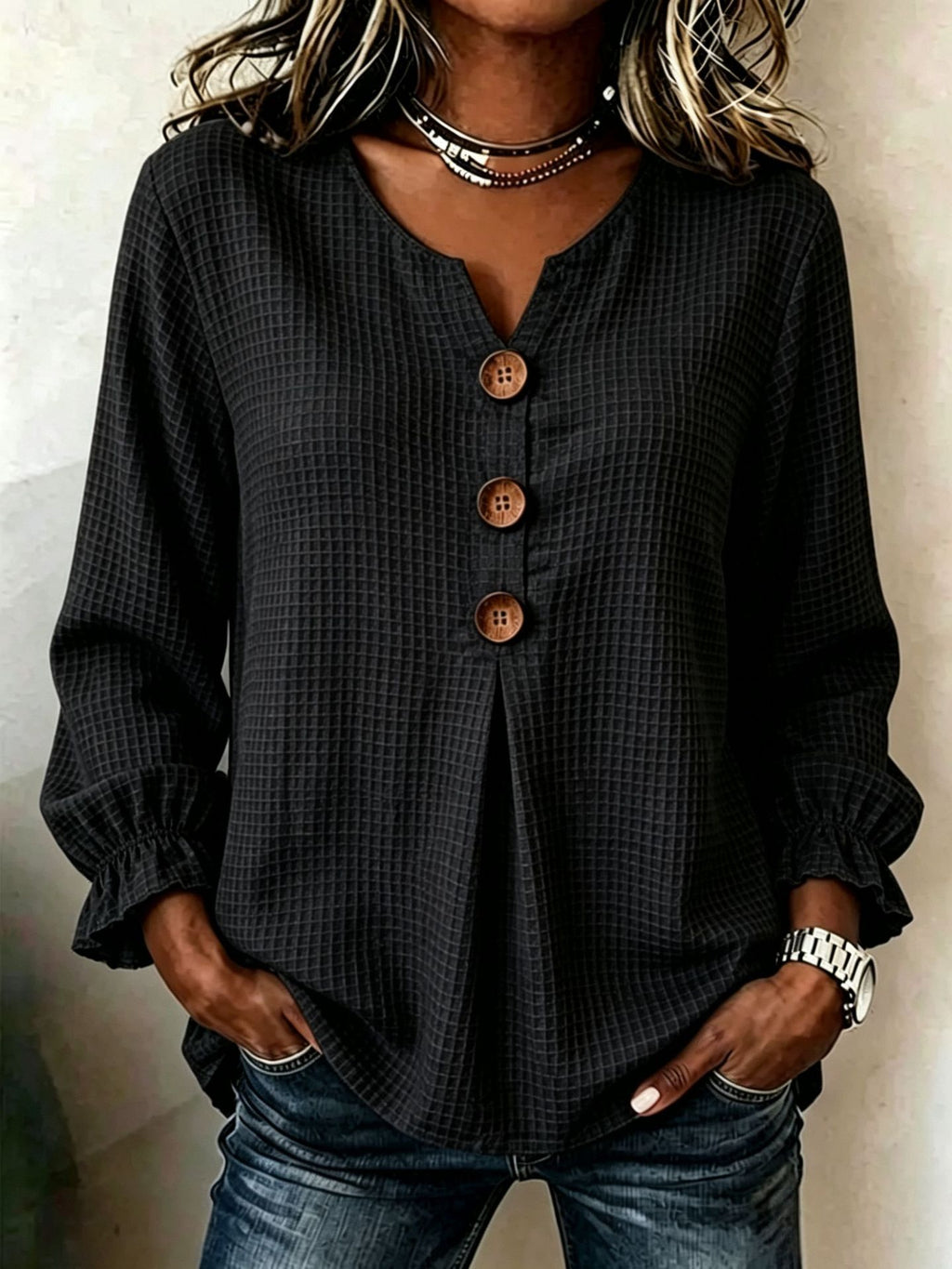A woman wearing a black long-sleeve button-up blouse with a subtle grid pattern and brown buttons, paired with blue jeans.