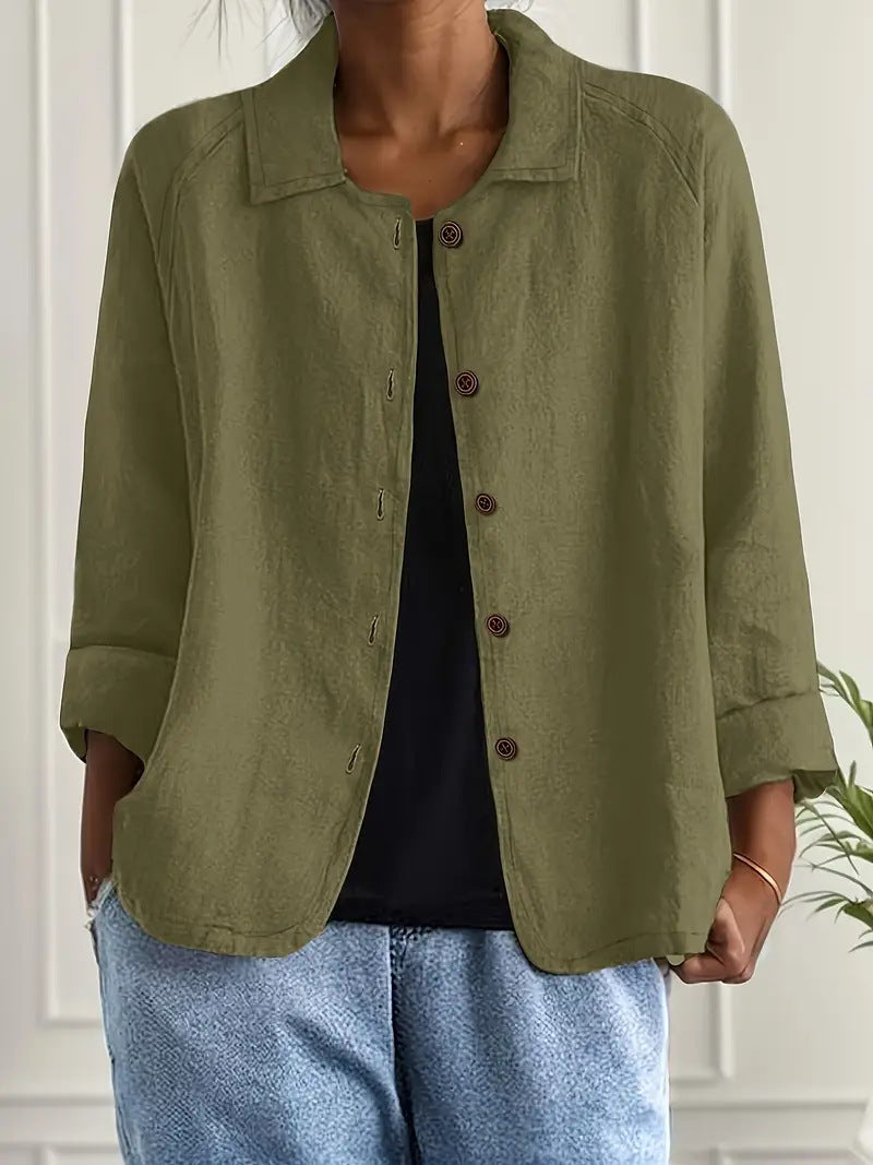 Person wearing an olive green jacket over a black top and blue jeans against a white wall.