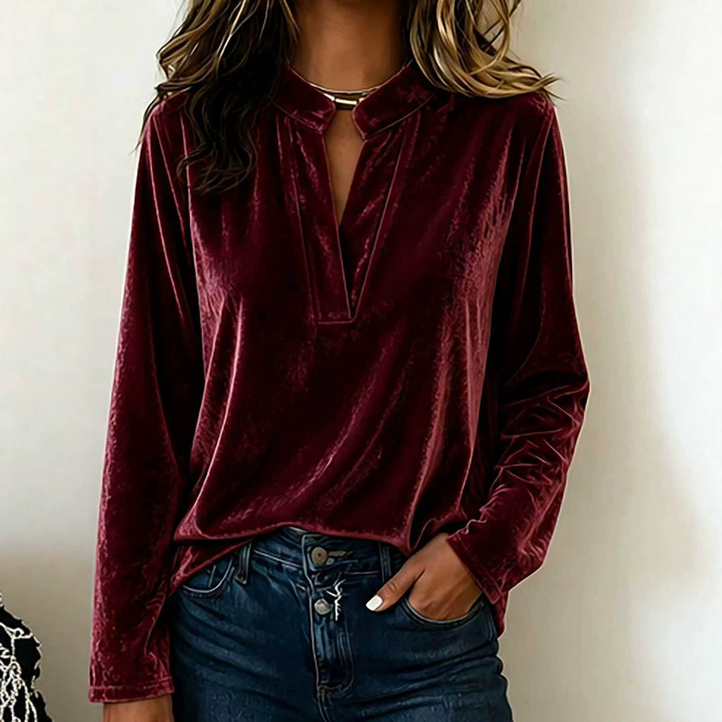 A woman wearing a long-sleeve burgundy velvet blouse with a split keyhole neckline, paired with dark blue jeans, shown against a plain off-white wall.