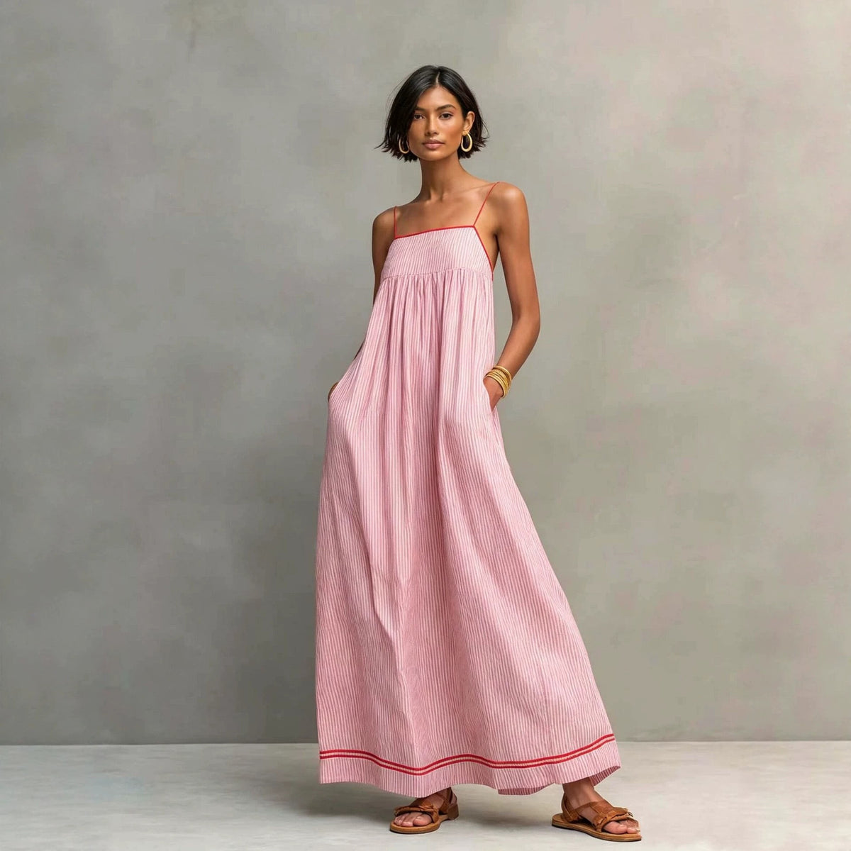 A woman modeling a light pink sleeveless maxi dress with thin shoulder straps, side pockets, and a subtle vertical stripe pattern, standing against a textured gray studio backdrop.