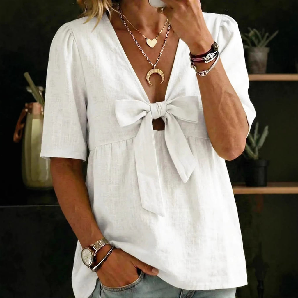Person wearing a white blouse with a bow detail, holding a phone, in a casual setting.