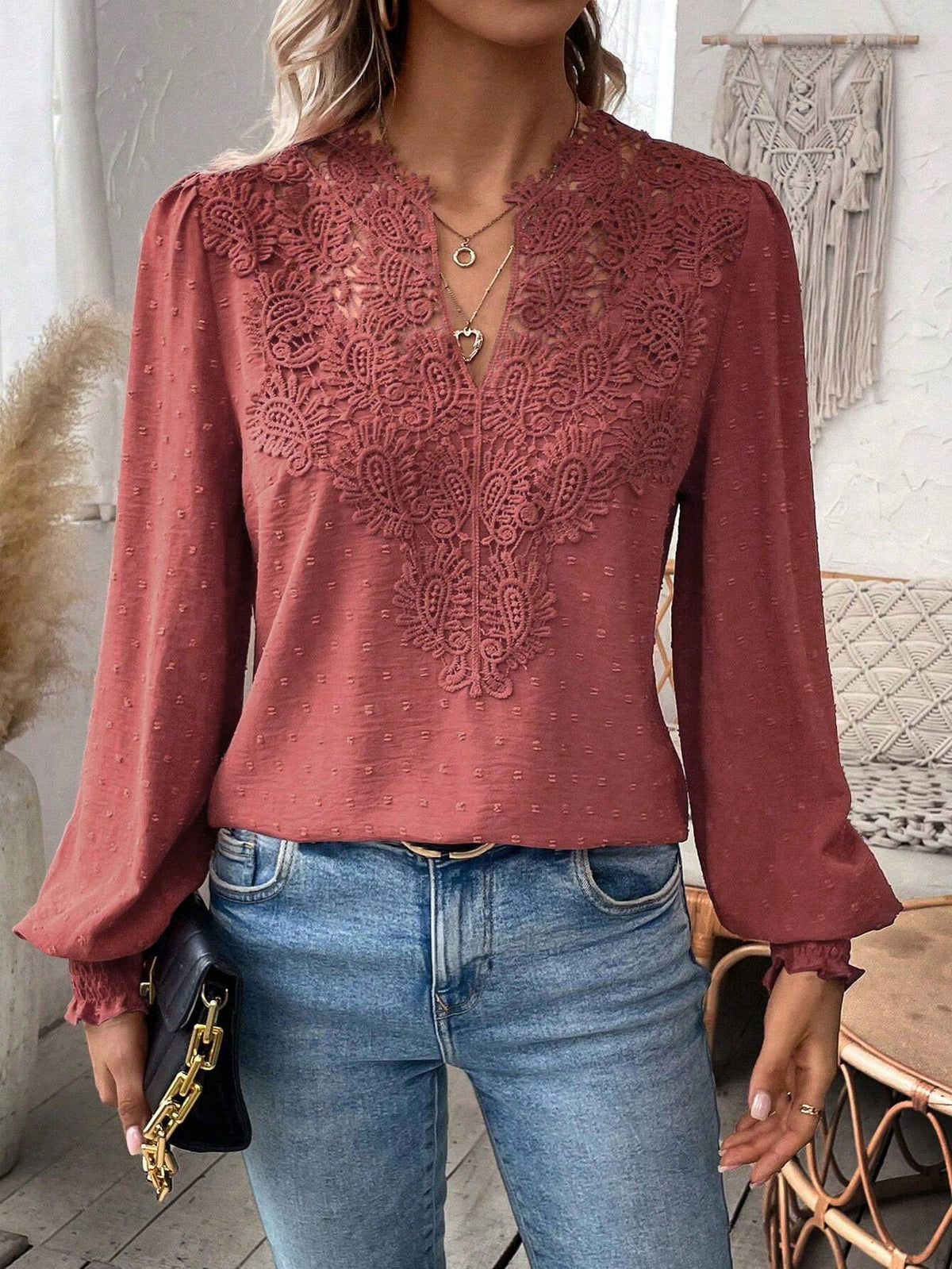 A woman wearing a dusty rose long-sleeve blouse with intricate lace applique on the chest, paired with blue jeans and holding a black clutch bag.