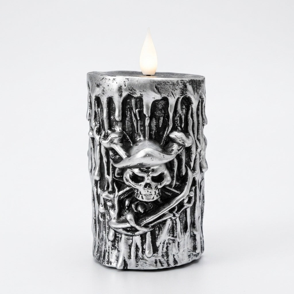 A metallic silver pillar candle with a textured, dripping wax finish and a raised skull and crossbones design on the front, shown lit with a small flame against a plain white background.