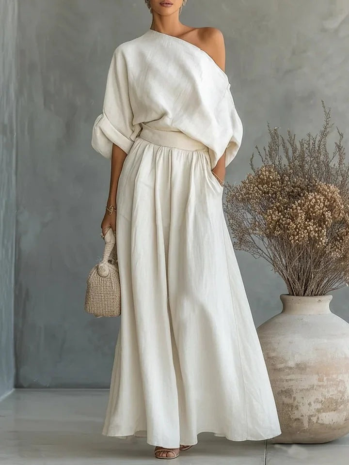 Woman wearing a white off-shoulder top and skirt standing against a gray wall with a vase of dried flowers.
