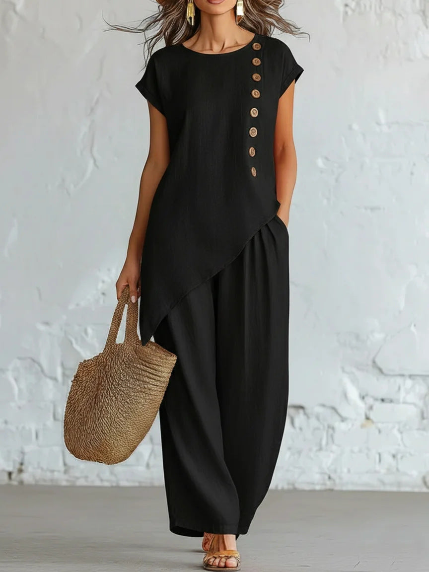 Woman wearing a black outfit with button details and holding a woven bag against a white brick wall.