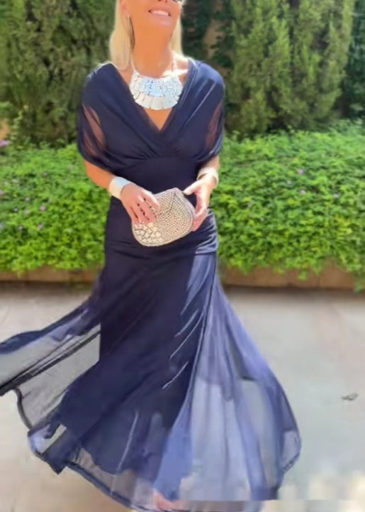 Woman in a navy blue evening gown holding a beaded clutch against a green hedge background
