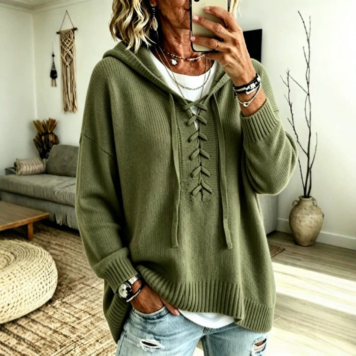 A woman takes a mirror selfie wearing an oversized olive green lace-up hooded sweater over a white undershirt and light-wash distressed jeans, in a bright living room setting.
