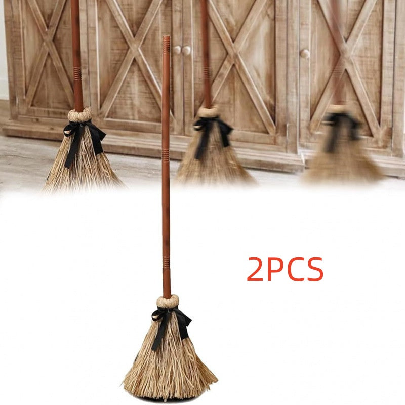 Three miniature straw brooms with brown handles and black ribbon bows, shown against a white foreground with a rustic wooden door in the background. Red text reads '3PCS'.
