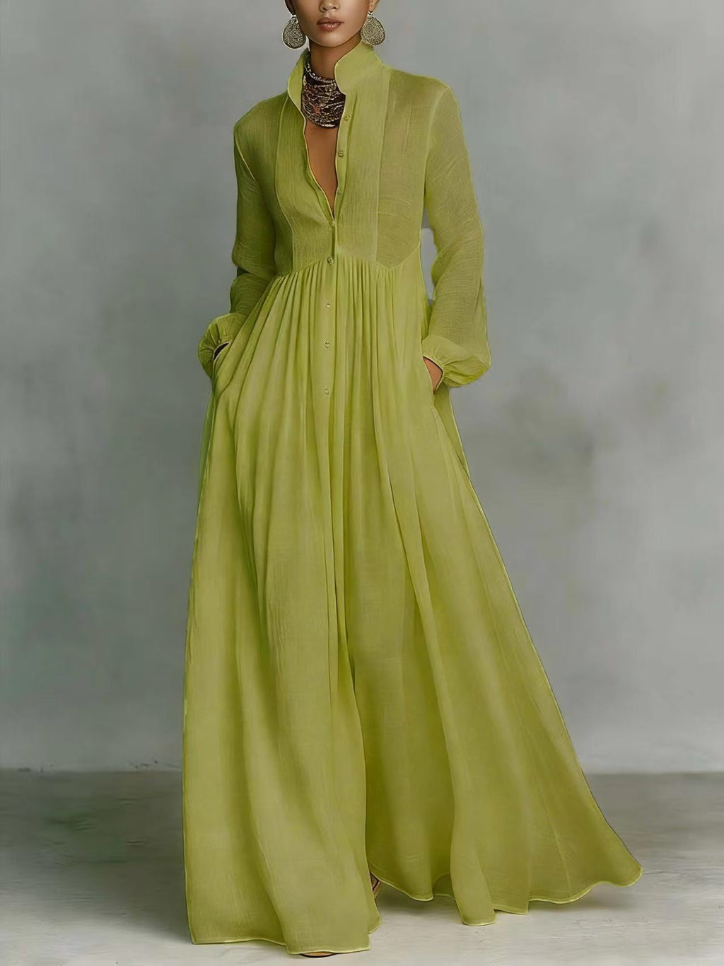 A woman models a flowing light green maxi dress with a deep V-neckline, long sleeves, and side pockets, set against a rustic white stucco wall with potted plants.