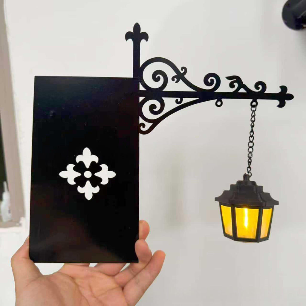 A hand holds a black decorative wall sign with a white floral motif, next to an ornate black metal bracket holding a hanging black lantern with a warm yellow light on a white wall.