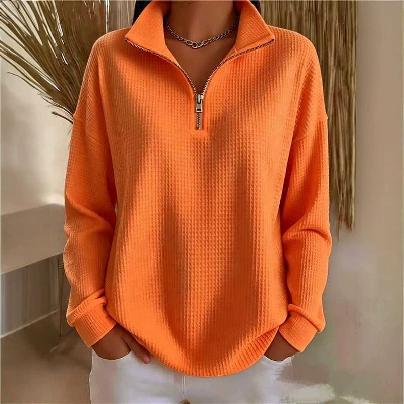 A person wearing a bright orange waffle-knit quarter-zip pullover sweater with a stand collar and silver zipper, paired with white pants and a silver chain necklace.