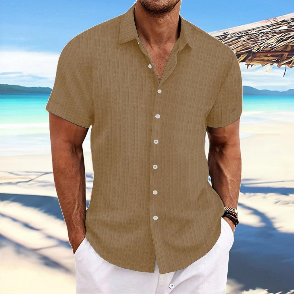 Man wearing a beige short-sleeve shirt on a beach with clear blue water and sky.