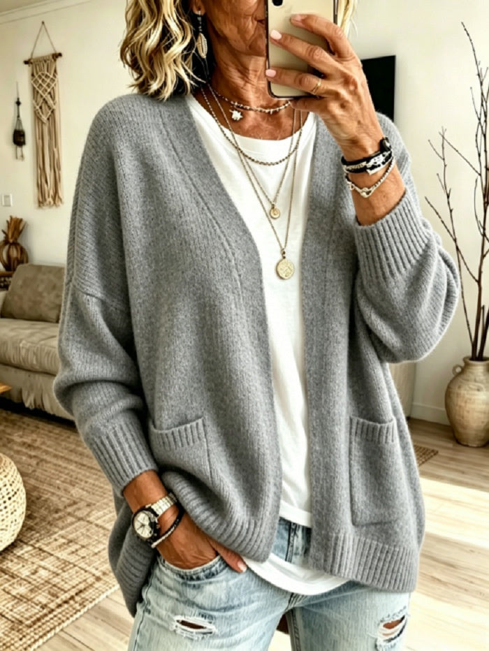 A woman takes a mirror selfie wearing a light gray knit cardigan over a white t-shirt and distressed light-wash jeans, accessorized with layered necklaces and bracelets in a cozy living room setting.