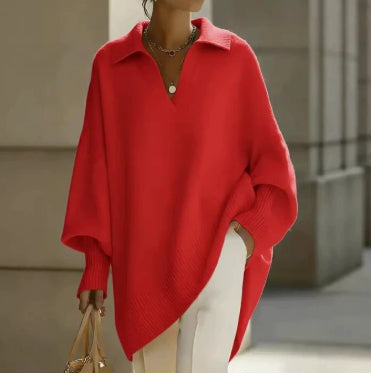 A woman wearing a bright red oversized knit polo sweater with wide draped sleeves and an asymmetrical hem, paired with white pants, standing in an outdoor architectural setting.